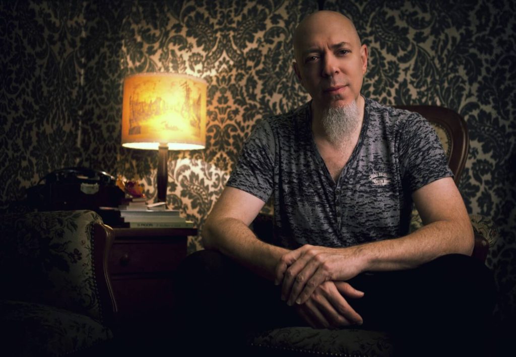 Jordan Rudess: man with grey/white goatee sitting down in a dark room with hands folded smiling in front of a lamp.